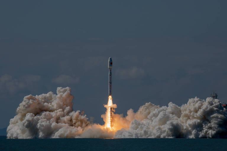 A imagem mostra um foguete sendo lançado a partir de uma plataforma sobre a água. O foguete está em ascensão, com chamas e fumaça saindo de seus motores, criando uma grande nuvem de vapor e fumaça ao redor da base. O céu ao fundo é azul, com algumas nuvens visíveis.