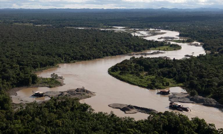 A imagem mostra uma vista aérea de um rio sinuoso que atravessa uma densa floresta. O rio apresenta águas de cor marrom e está cercado por vegetação verdejante. Em algumas áreas do rio, há pequenas ilhas de areia e embarcações visíveis. O céu está parcialmente nublado, e a paisagem se estende até o horizonte, onde se podem ver montanhas ao fundo.