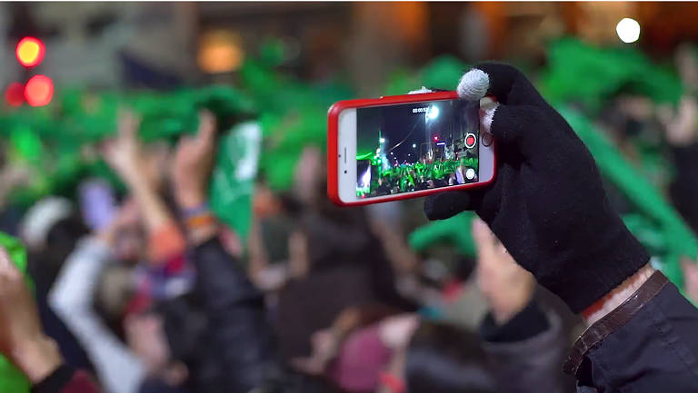 Uma mão segura um celular que está filmando uma manifestação.