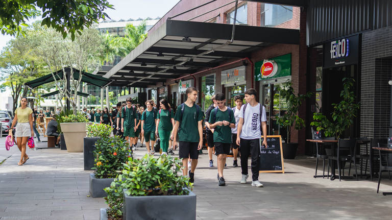 A imagem mostra um grupo de estudantes vestidos com uniformes verdes caminhando em uma área comercial. Eles estão na calçada, cercados por lojas e vegetação. À esquerda, uma mulher carrega sacolas de compras. O ambiente é ensolarado e moderno, com mesas e cadeiras visíveis em algumas das lojas.