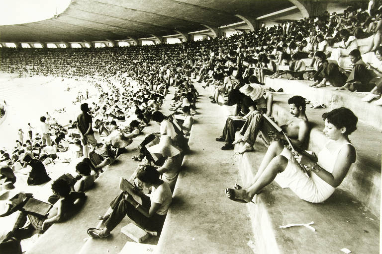 A imagem mostra um grande público em um estádio, com muitas pessoas sentadas nas arquibancadas. Algumas pessoas estão segurando objetos, possivelmente instrumentos ou equipamentos de gravação. O ambiente é de uma época anterior, com uma estética em preto e branco, sugerindo um evento esportivo ou cultural. O estádio está lotado, e a atmosfera parece animada.