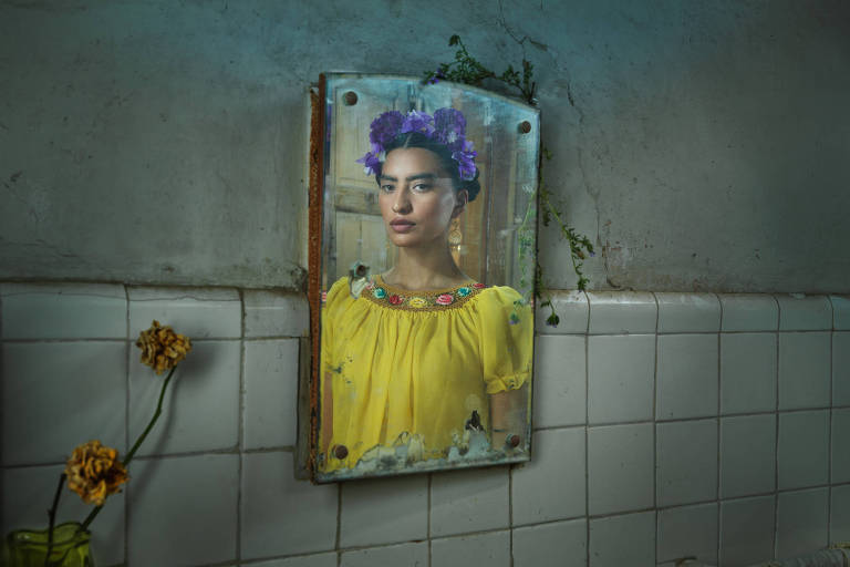 A imagem mostra um espelho antigo em uma parede com azulejos brancos. No reflexo, uma mulher com um vestido amarelo e uma coroa de flores roxas aparece. O ambiente é rústico, com paredes desgastadas e um vaso com flores secas ao lado do espelho. Há também algumas plantas verdes crescendo nas bordas do espelho.