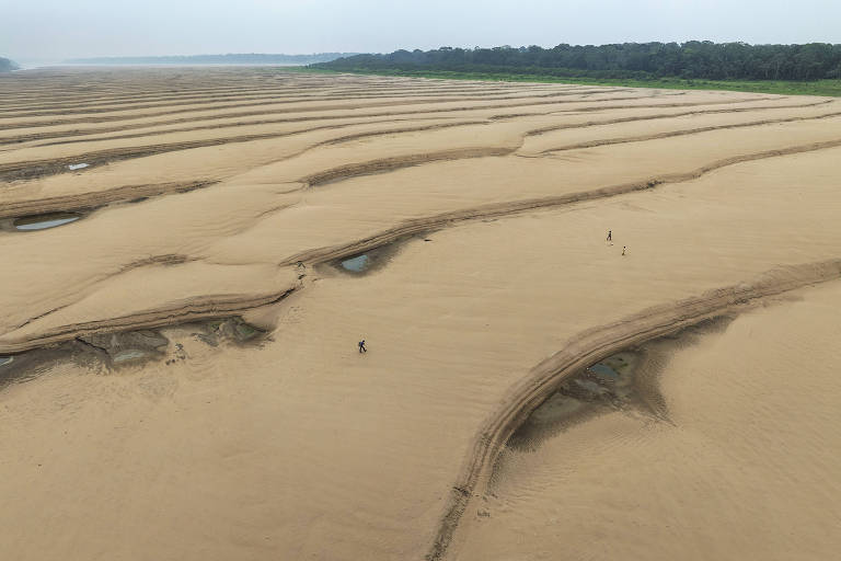  imagem mostra uma vasta área de areia com ondulações e pequenas poças de água. Ao fundo, há uma vegetação densa, possivelmente uma floresta. A cena é ampla e parece ter sido capturada de uma altura considerável, revelando a extensão da areia e a disposição das poças. Algumas figuras humanas são visíveis, caminhando sobre a areia.