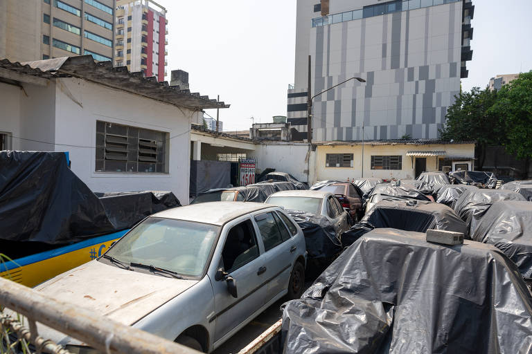 A imagem mostra um estacionamento com vários carros cobertos por lonas. Ao fundo, há um edifício alto e moderno, enquanto à esquerda se vê uma construção mais baixa com janelas. O ambiente parece ser urbano, com outros prédios visíveis ao fundo.