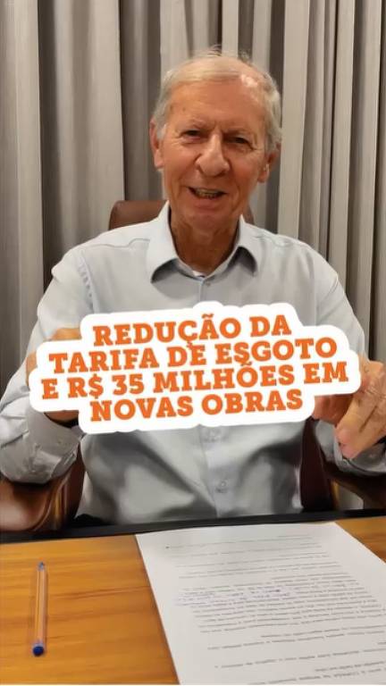 Um homem sorridente, com cabelo grisalho, está sentado em uma mesa. Ele segura um cartaz que diz 'REDUÇÃO DA TARIFA DE ESGOTO E R$ 35 MILHÕES EM NOVAS OBRAS'. Ao fundo, há cortinas e uma folha de papel sobre a mesa.