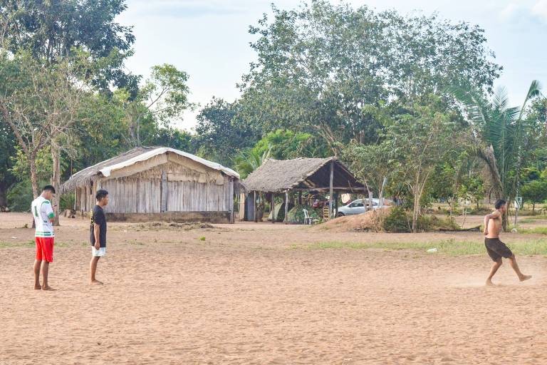 A imagem mostra três jovens em uma área de terra batida, com duas casas de madeira ao fundo. Dois jovens estão parados, um deles olhando para o celular, enquanto um terceiro jovem corre em direção a eles. A vegetação ao redor é densa, com árvores e plantas típicas da região. O céu está parcialmente nublado.