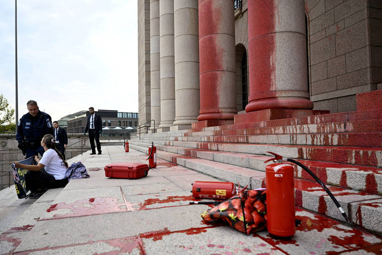 A imagem mostra escadas de um edifício com colunas, onde há manchas vermelhas no chão, sugerindo um ato de protesto. Dois extintores de incêndio estão visíveis, um deles derramado. Uma mulher está sentada no degrau, enquanto duas pessoas em trajes formais estão ao fundo, observando a cena.