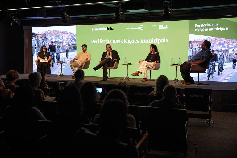 A imagem mostra um painel de debate com cinco pessoas sentadas em cadeiras em um palco. Ao fundo, há uma tela verde com o texto 'Periferias nas eleições municipais'. O ambiente é moderno, com uma plateia visível na parte inferior da imagem. As pessoas no painel estão vestidas de maneira casual e parecem estar discutindo um tema relacionado às eleições