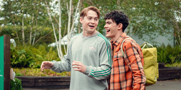 A imagem mostra dois jovens sorrindo e conversando ao ar livre. Um deles está vestindo um moletom cinza com listras verdes e o outro está usando uma camisa xadrez laranja. O jovem com o moletom parece estar gesticulando enquanto fala. Ao fundo, há árvores e uma vegetação verde, sugerindo um ambiente natural.