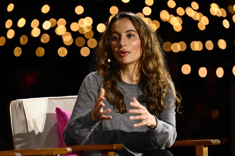 A imagem mostra uma mulher com cabelo longo e ondulado， usando uma blusa cinza， sentada em uma cadeira. Ela está gesticulando enquanto fala， com um fundo de luzes desfocadas. A atmosfera é acolhedora e informal.
