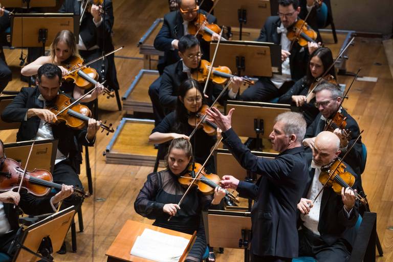 A imagem mostra uma orquestra em performance, com músicos tocando violinos e outros instrumentos. O maestro está em destaque, gesticulando enquanto conduz a apresentação. Os músicos estão organizados em fileiras, com um fundo de palco de madeira.