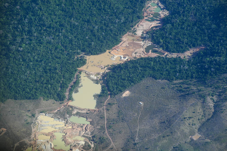 A imagem mostra uma vista aérea de uma área com desmatamento, onde se pode observar uma mistura de solo exposto e áreas de vegetação densa. Há também corpos d'água em diferentes tonalidades, indicando possíveis lagos ou poços. A paisagem é marcada por uma transição entre áreas desmatadas e florestas.