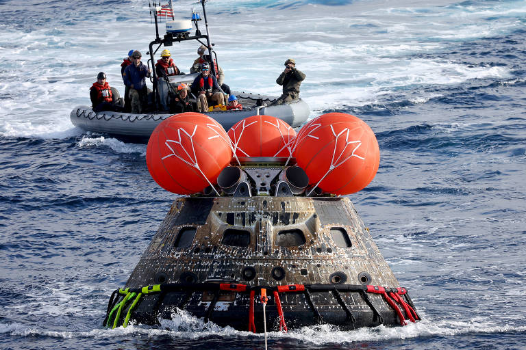 A imagem mostra uma nave espacial com três grandes balões laranja flutuando no mar. Ao fundo, há uma embarcação de resgate com várias pessoas a bordo, algumas delas usando coletes salva-vidas. O mar está agitado, e há ondas visíveis ao redor da nave.