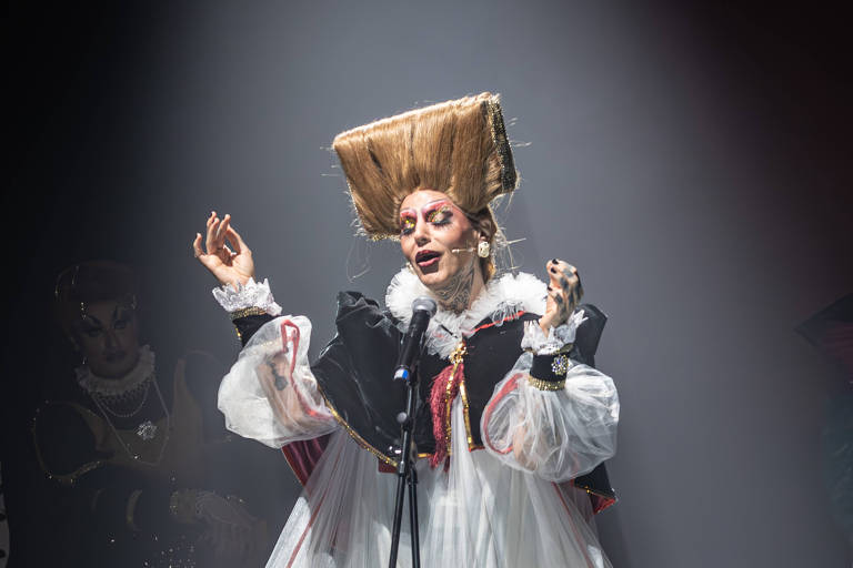 A imagem mostra uma pessoa em uma apresentação teatral, vestindo um vestido longo e volumoso, predominantemente branco, com detalhes escuros. A figura está em um palco iluminado por um feixe de luz, com as mãos levantadas em uma pose expressiva. Ao fundo, é possível ver silhuetas de outras pessoas, sugerindo uma plateia ou outros participantes da performance.