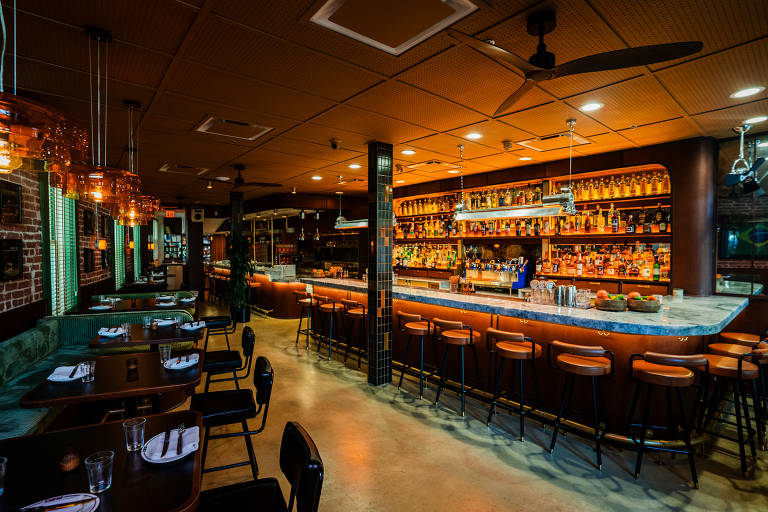 A imagem mostra o interior de um restaurante moderno, com um bar ao fundo. O bar é iluminado por luzes quentes e exibe uma variedade de garrafas dispostas em prateleiras. As mesas estão organizadas na parte frontal, com cadeiras pretas e toalhas de mesa brancas. O ambiente é acolhedor, com um piso de concreto e paredes decoradas. Há também ventiladores de teto visíveis.