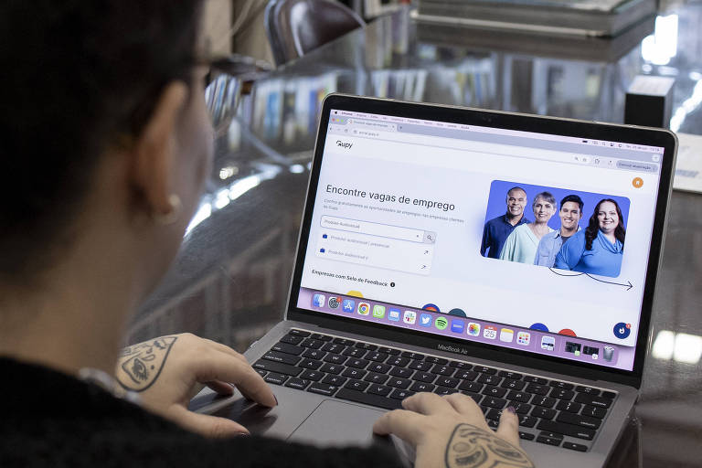 A imagem mostra uma pessoa de costas, com cabelo cacheado preso em um coque, usando um laptop em uma mesa em uma biblioteca. Ao fundo, há estantes com livros organizados. Na tela do laptop, é visível um site com a frase 'Encontre vagas de emprego' e imagens de quatro pessoas. A mesa é de madeira e reflete a luz ambiente.