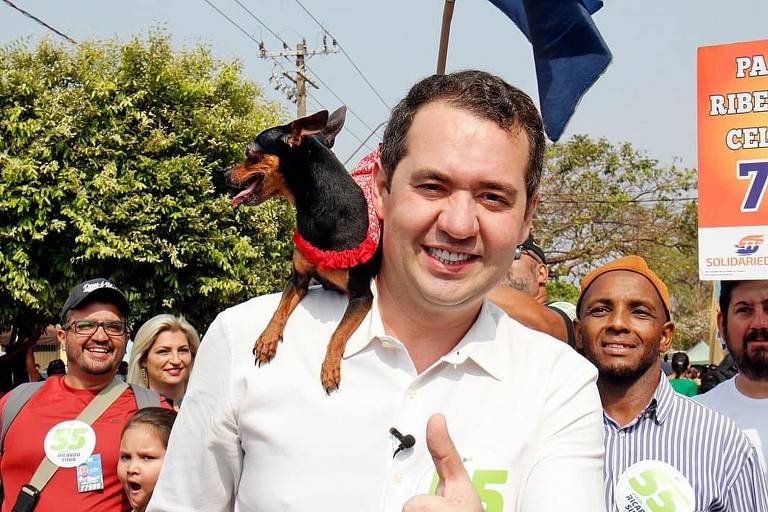 Imagem mostra um homem sorrindo, fazendo sinal de positivo com a mão esquerda; em seu ombro direito, há um pequeno cão