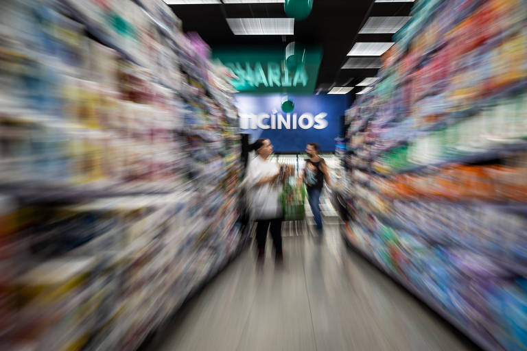 A imagem mostra um corredor de supermercado com prateleiras cheias de produtos. O foco está desfocado, mas é possível ver duas pessoas em movimento no corredor. Ao fundo, há uma placa com a palavra 'Ofertas' visível. O ambiente é bem iluminado.