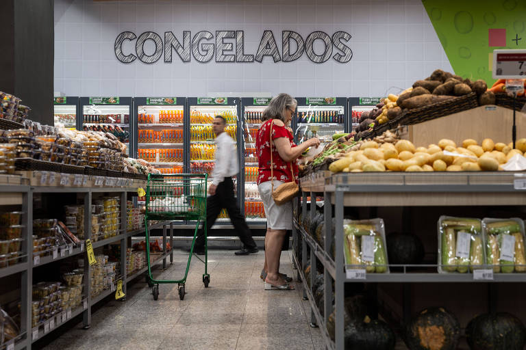 Imagem mostra uma mulher escolhendo batatas em uma banca de legumes no supermercado. Um homem passa ao fundo e acima dele há uma placa onde está escrito: Congelados