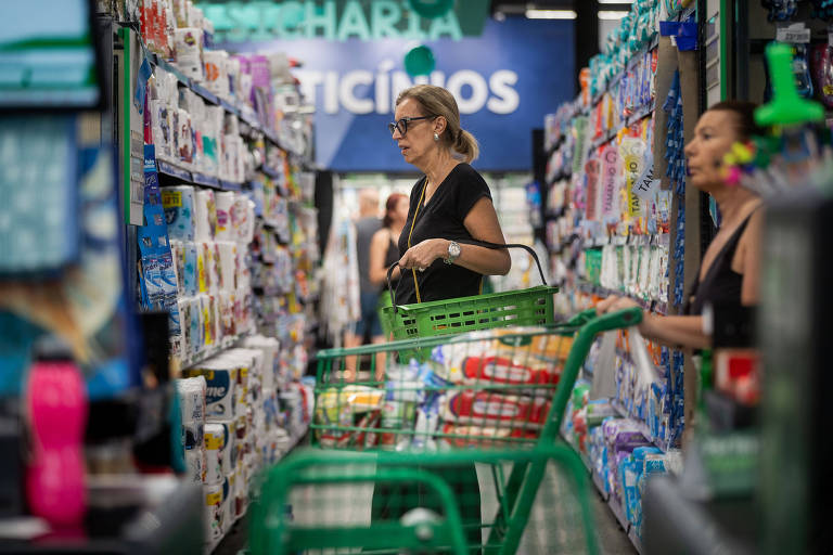 A imagem mostra uma mulher com um carrinho de compras verde em um corredor de supermercado. Ela está olhando para os produtos nas prateleiras， que estão cheias de itens variados. Ao fundo， há outra mulher também fazendo compras. O ambiente é bem iluminado e organizado.