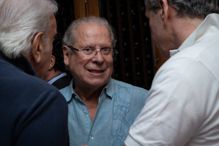 A imagem mostra um grupo de homens conversando em um ambiente interno. Um homem de camisa azul clara está sorrindo e olhando para os outros， enquanto dois homens， um com cabelo grisalho e outro com cabelo claro， estão parcialmente visíveis ao fundo. O ambiente parece ser acolhedor， com uma parede de madeira ao fundo.