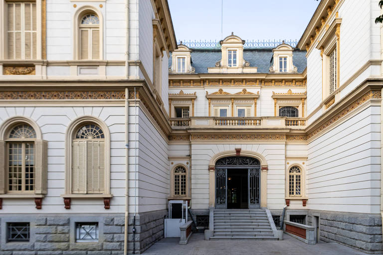 A imagem mostra a fachada de um edifício histórico， com paredes bege claro e detalhes em dourado. A entrada principal é composta por escadas que levam a um grande portal central， flanqueado por janelas com persianas. O telhado é inclinado e possui janelas no sótão. O céu está claro， indicando um dia ensolarado.