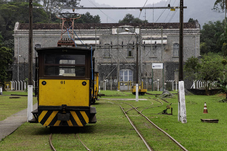 A imagem mostra um trem amarelo e preto em primeiro plano, com o número 01 visível. Ao fundo, há um edifício de pedra cinza com janelas grandes e uma estrutura de telhado danificada. A área ao redor é verde, com grama e algumas árvores. Trilhos de trem se estendem a partir do trem em direção ao edifício. Há postes de eletricidade e uma sinalização visível.