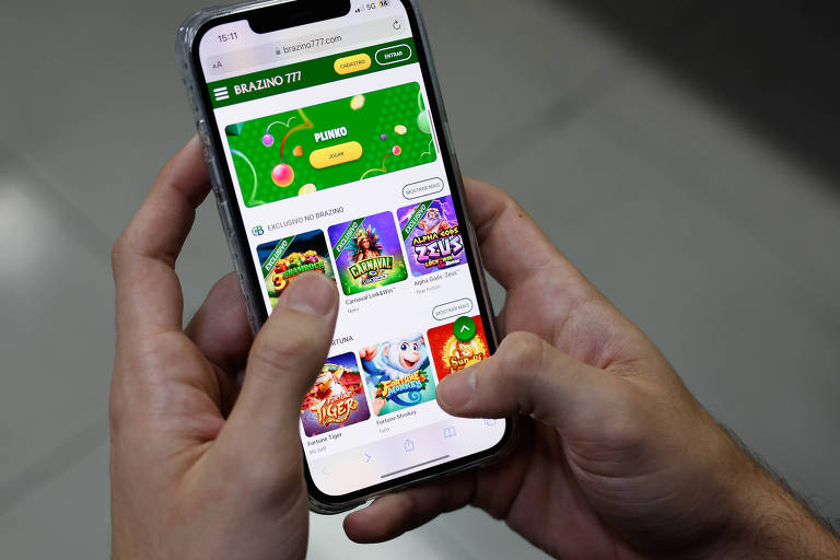 Uma pessoa está segurando um smartphone com ambas as mãos. A tela do dispositivo exibe uma interface de aplicativo com várias opções de jogos, incluindo gráficos coloridos e ícones. O fundo é de piso claro e a iluminação é suave.