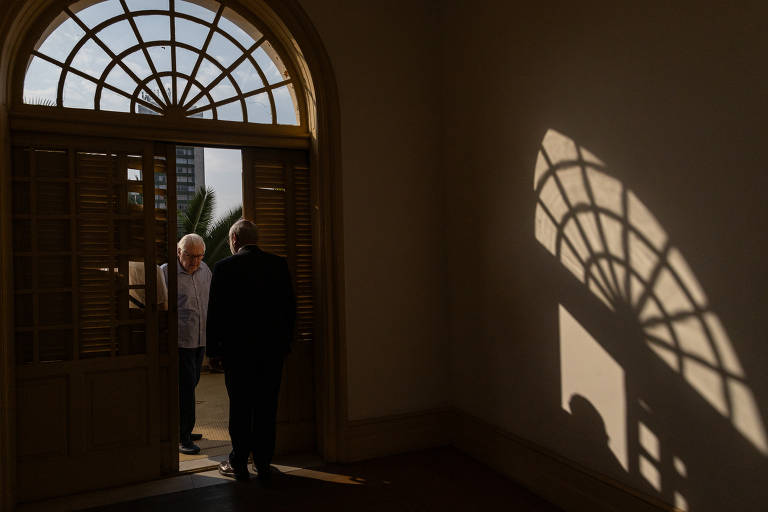 A imagem mostra duas pessoas em uma porta aberta, com luz natural entrando. Uma pessoa está de costas, vestindo um terno escuro, enquanto a outra, de cabelo claro, está de frente, usando uma camisa clara. A luz que atravessa a porta entreaberta e um vitral em formato de semicírculo acima da porta se projeta numa parede escura do interior de um cômodo.