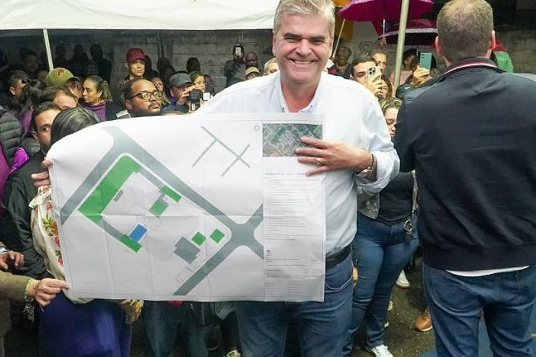 A imagem mostra um homem sorridente segurando um grande mapa em formato de folha, que parece representar um plano de área. Ele está em um evento ao ar livre, cercado por uma multidão de pessoas que observam. Algumas pessoas estão sorrindo e outras parecem atentas ao que está acontecendo. O ambiente é festivo, com tendas ao fundo.