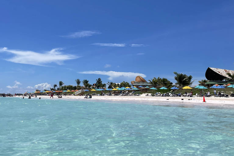 A imagem mostra uma praia com água cristalina e um céu azul claro. Na areia, há várias pessoas, algumas deitadas e outras caminhando. Ao fundo, há uma área com cadeiras e guarda-sóis coloridos, além de palmeiras. O ambiente é ensolarado e tranquilo.