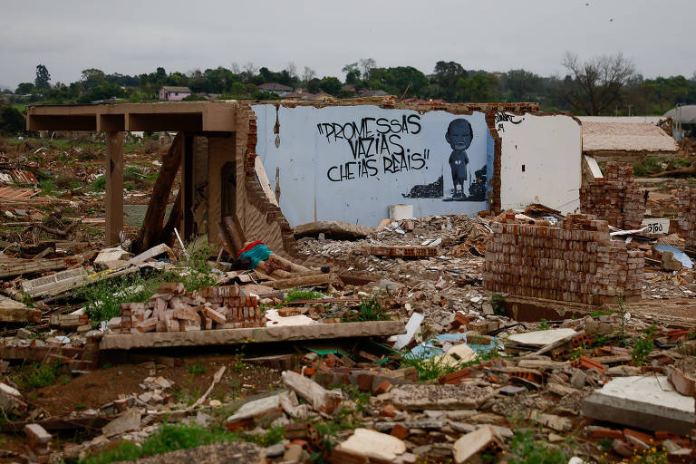 A imagem mostra uma área devastada， com destroços de uma construção e um mural na parede de um edifício parcialmente destruído. O chão está coberto de entulho e restos de materiais de construção， com vegetação ao fundo.
