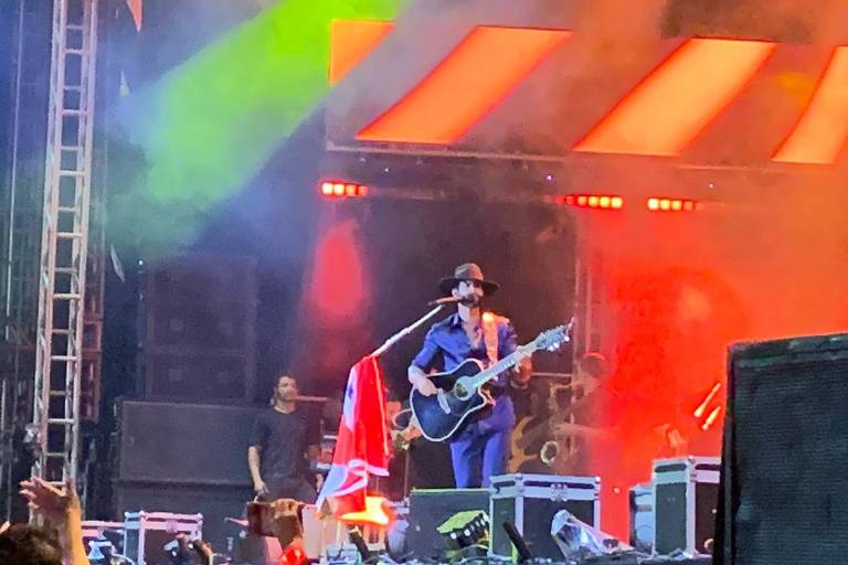 Um músico se apresenta em um palco iluminado com luzes coloridas. Ele está vestido com uma camisa azul e um chapéu, tocando um violão. Atrás dele, há uma bandeira pendurada e um fundo com luzes vermelhas e verdes. Um membro da equipe pode ser visto ao fundo, e a plateia está visível na parte inferior da imagem.