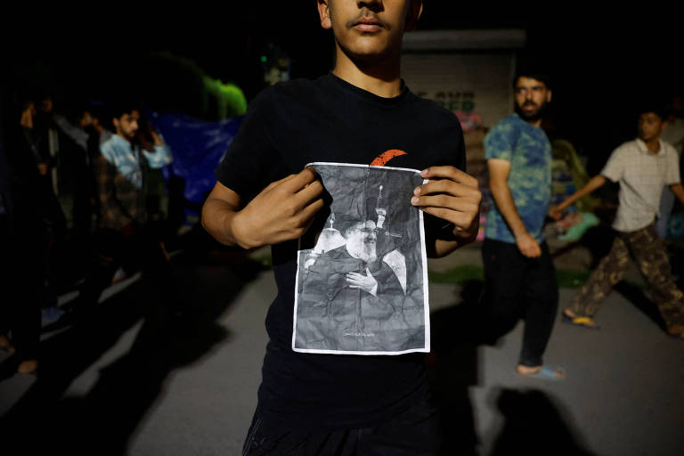 Imagem mostra movimentação de pessoas a noite em uma rua; o menino ao centro tem meio rosto na foto, usa camiseta preta, e segura imagem do líder morto.