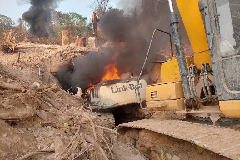 A imagem mostra uma escavadeira, da marca Link-Belt, pegando fogo em uma área de desmatamento. Há fumaça preta saindo da máquina e do solo ao redor, que está coberto de terra e troncos de árvores derrubadas. O ambiente é árido, com vegetação escassa ao fundo.