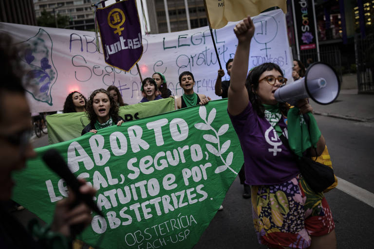 A imagem mostra um grupo de pessoas participando de uma manifestação nas ruas. Elas seguram faixas e cartazes, com destaque para um grande banner verde que diz 'ABORTO LEGAL, SEGURO, GRATUITO E POR OBSTETRIZES'. Algumas manifestantes estão usando lenços verdes e uma delas está falando ao microfone. Ao fundo, há outros cartazes e uma atmosfera de protesto.