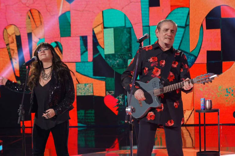 A imagem mostra dois artistas se apresentando em um palco. À esquerda, uma mulher com cabelo longo e solto, vestindo uma blusa preta brilhante e calças pretas, está cantando em um microfone. À direita, um homem mais velho, usando uma camisa preta com flores vermelhas e calças escuras, toca um violão de cor cinza. O fundo é colorido, com formas geométricas e corações em tons vibrantes de rosa, verde e azul. Há também um suporte com um copo ao fundo.