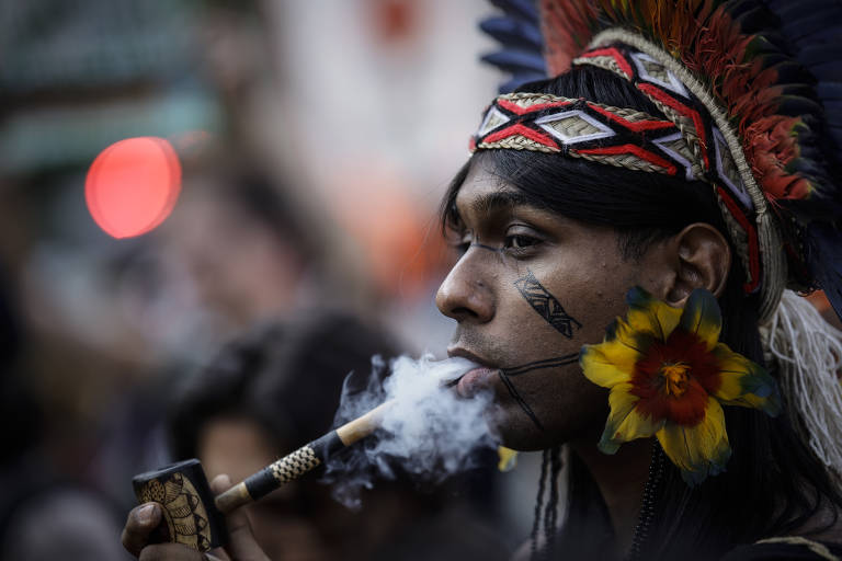 A imagem mostra um homem indígena usando um cocar colorido e segurando um cachimbo. Ele está soprando fumaça, e há flores presas em sua orelha. O fundo é desfocado, com luzes e pessoas indistintas.