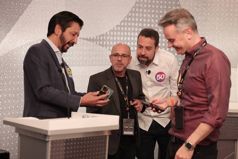 Quatro homens estão reunidos em um evento, olhando para um celular que um deles está segurando. O homem à esquerda, com cabelo escuro e barba, usa um paletó escuro e uma camiseta branca. O segundo homem, de óculos e cabelo curto, está no centro, vestindo uma camisa preta. O terceiro homem, com cabelo grisalho e barba, usa uma camisa branca e um crachá com o número 50. O quarto homem, à direita, tem cabelo curto e usa uma camisa marrom. Todos parecem concentrados no celular.