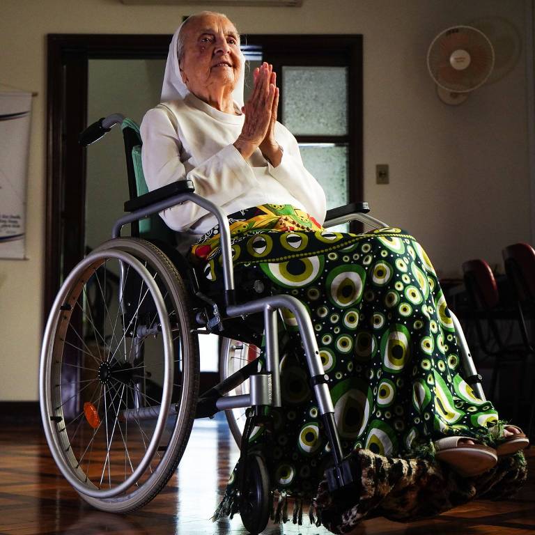 Uma mulher idosa está sentada em uma cadeira de rodas, com as mãos unidas em um gesto de oração ou agradecimento. Ela usa um vestido longo com estampas circulares em verde e preto e um lenço na cabeça. O ambiente ao fundo é iluminado, com uma porta aberta e uma parede clara.