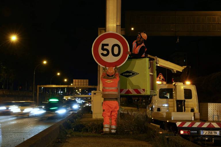 A imagem mostra dois trabalhadores em um canteiro de obras à noite. Um deles está em uma plataforma elevatória, instalando uma placa de limite de velocidade de 50 km/h, enquanto o outro segura a placa. Ao fundo, há um tráfego de veículos com faróis acesos e uma iluminação pública visível.