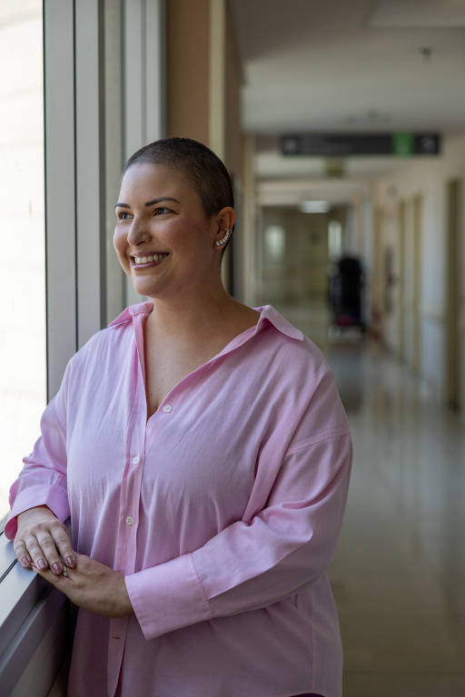 Mulher branca de cabelo raspado está em pé em frente a uma janela. Ela usa uma blusa social rosa