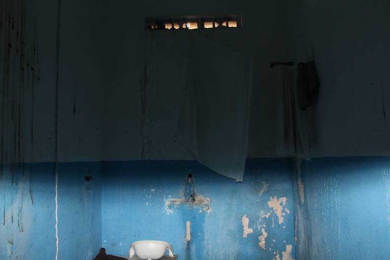 A imagem mostra o interior de uma cela com paredes pintadas de azul. Há uma janela pequena no alto, permitindo a entrada de luz. Um vaso sanitário branco está visível no canto inferior direito. Algumas toalhas brancas estão penduradas em um suporte na parede. As paredes apresentam marcas de desgaste e sujeira, com manchas e descascados visíveis.