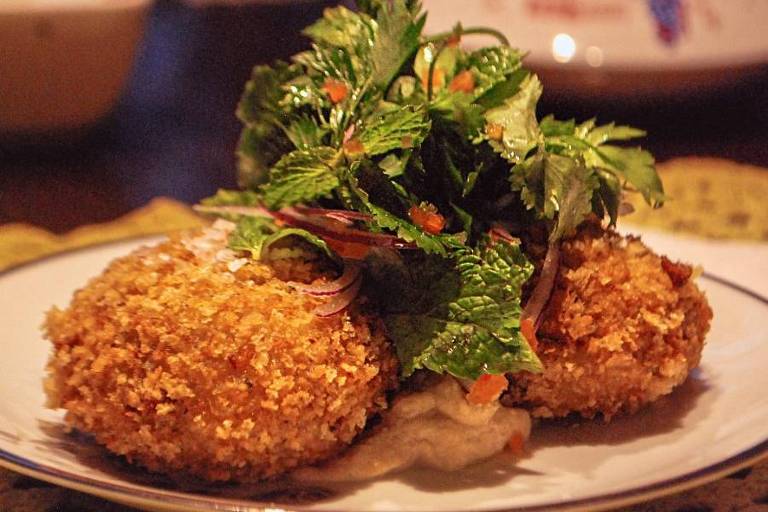 A imagem mostra um prato com dois croquetes empanados e fritos, cobertos com uma mistura de ervas frescas, incluindo folhas verdes e tiras de cebola roxa. A base do prato parece ter um molho claro, possivelmente de tahine ou similar. O fundo é desfocado, mas sugere um ambiente de refeição.