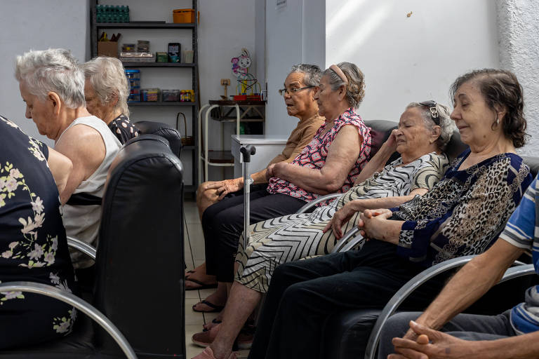 A imagem mostra um grupo de idosos sentados em cadeiras em um ambiente interno. Eles estão em uma sala com paredes claras e prateleiras ao fundo, onde há alguns objetos visíveis. Os idosos estão vestidos de maneira casual, com algumas mulheres usando blusas estampadas. A maioria parece estar atenta ou relaxada, com expressões serenas. Um dos idosos usa um andador. A iluminação é natural, entrando pela janela.