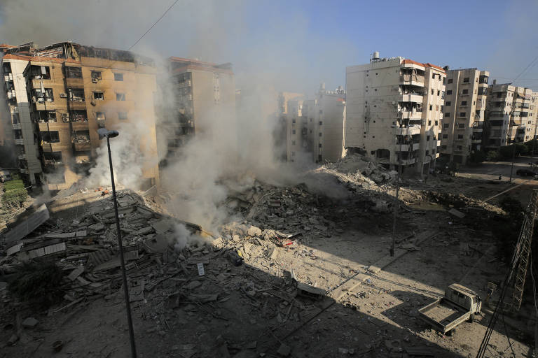 A imagem mostra uma área urbana com prédios ao fundo e uma grande quantidade de fumaça saindo de uma área em ruínas, onde um edifício foi destruído. O chão está coberto de escombros e detritos, e a cena é iluminada pela luz do dia.