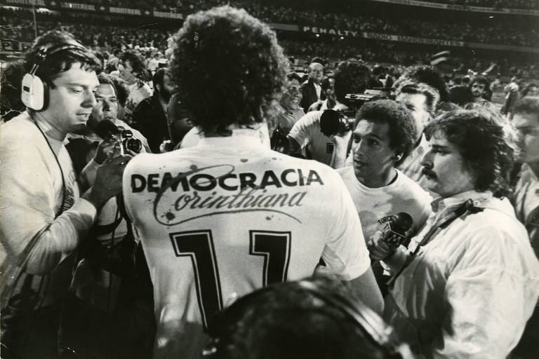 Fotografia em preto e branco mostra jogadores aglomerados em comemoração no centro de um estádio de futebol. Eles usam camisetas que dizem "democracia corinthiana".