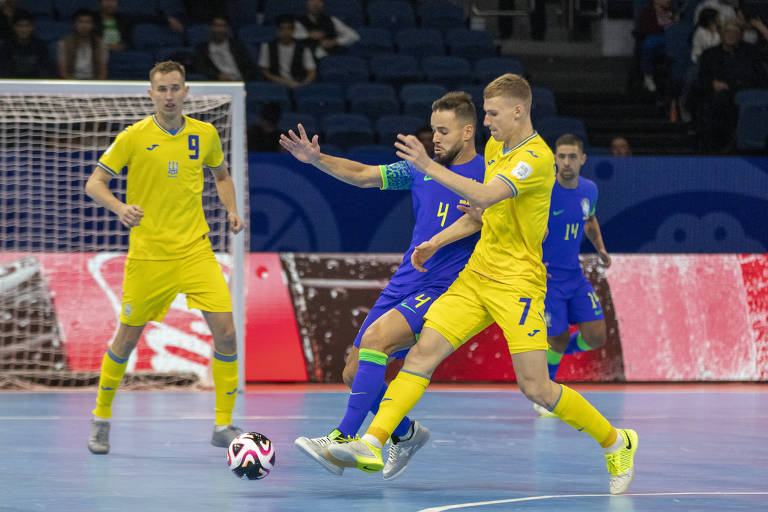 A imagem mostra uma partida de futsal entre jogadores do Brasil e da Ucrânia. Um jogador brasileiro, vestido com uniforme azul e número 4, está em disputa de bola com um jogador ucraniano, que usa um uniforme amarelo com o número 9. Outros jogadores ucranianos estão visíveis ao fundo, e a rede do gol é parcialmente visível. O ambiente é de um ginásio esportivo, com espectadores ao fundo.