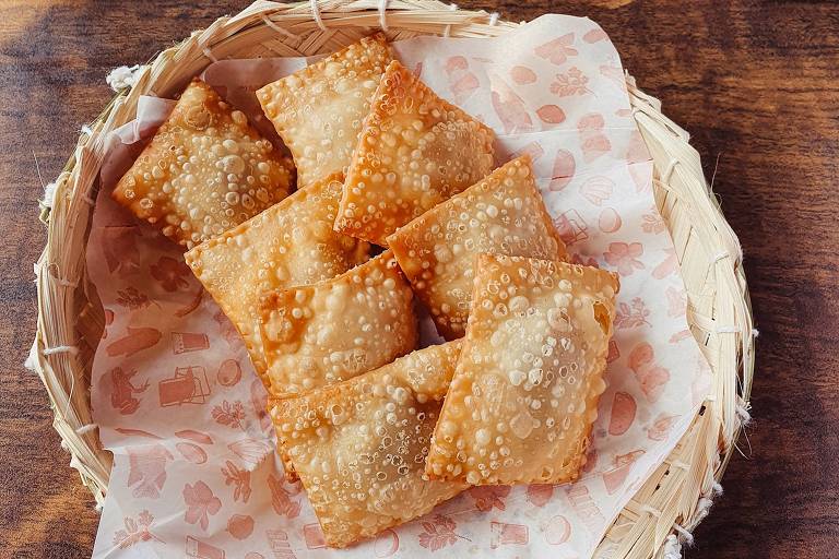 A imagem mostra uma cesta de vime com pastéis fritos dispostos sobre um papel decorado. Ao lado, há um pequeno recipiente de pedra com guacamole e uma garrafinha de molho laranja. Ao fundo, três pratos azuis empilhados sobre uma mesa de madeira.