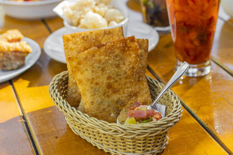 A imagem mostra uma cesta de vime com três salgados fritos, que parecem ser pastéis, acompanhados de um pequeno recipiente com uma mistura de legumes picados, incluindo tomate e pimentão. Ao fundo, há outros pratos e uma bebida em um copo transparente.